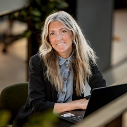 Kvinne med langt hår og svart jakke sitter ved en laptop og smiler.