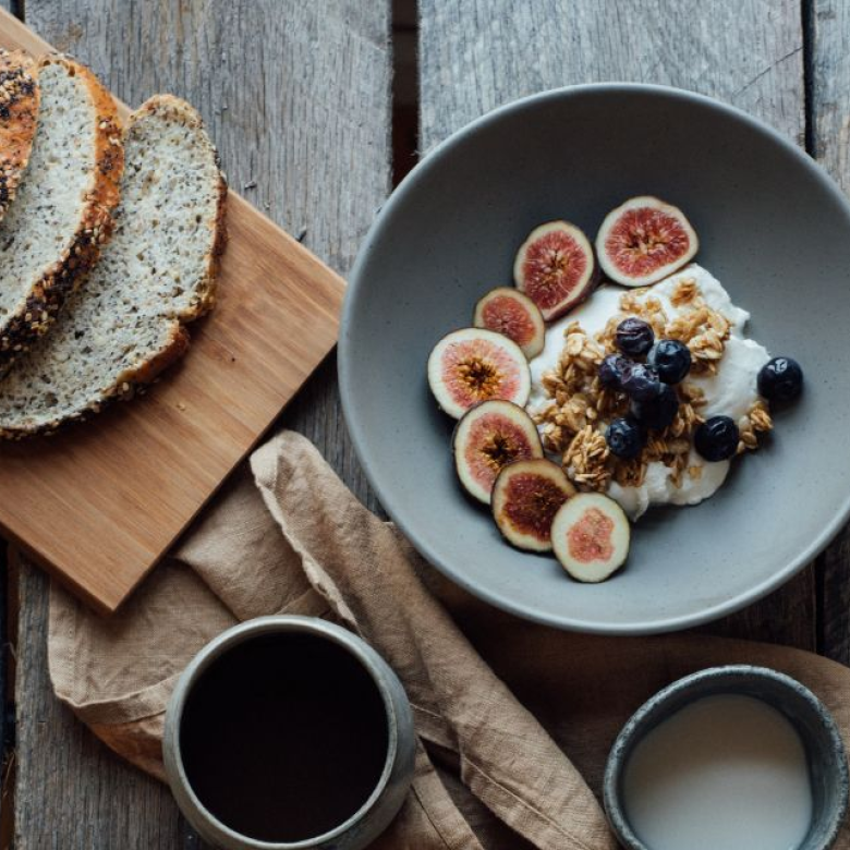 Frokost med brødskiver, yoghurt med frukt og nøtter, kaffe og melk på trebord.