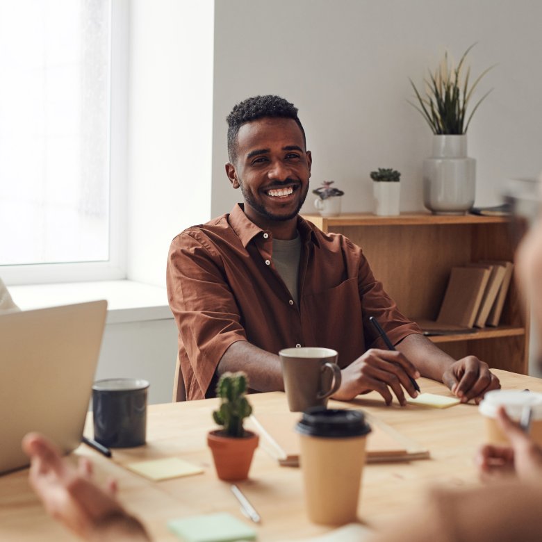 Tre personer smiler og diskuterer rundt et bord med bærbar PC og kaffekopper.