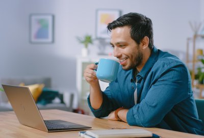 Mann drikker kaffe mens han jobber på en bærbar datamaskin hjemme.