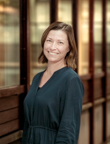 Kvinne i blå kjole smiler foran en bygning med store vinduer.